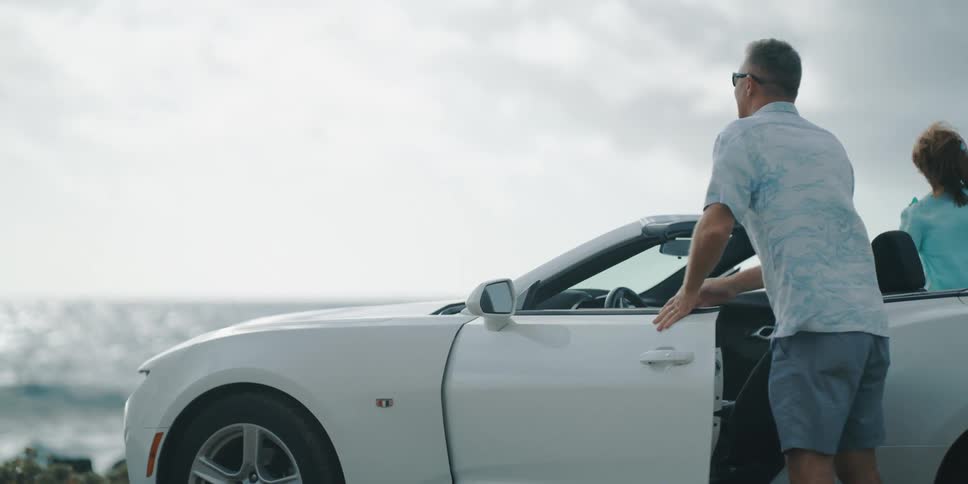 Older couple parks white convertible near ocean and walks towards ocean holding hands