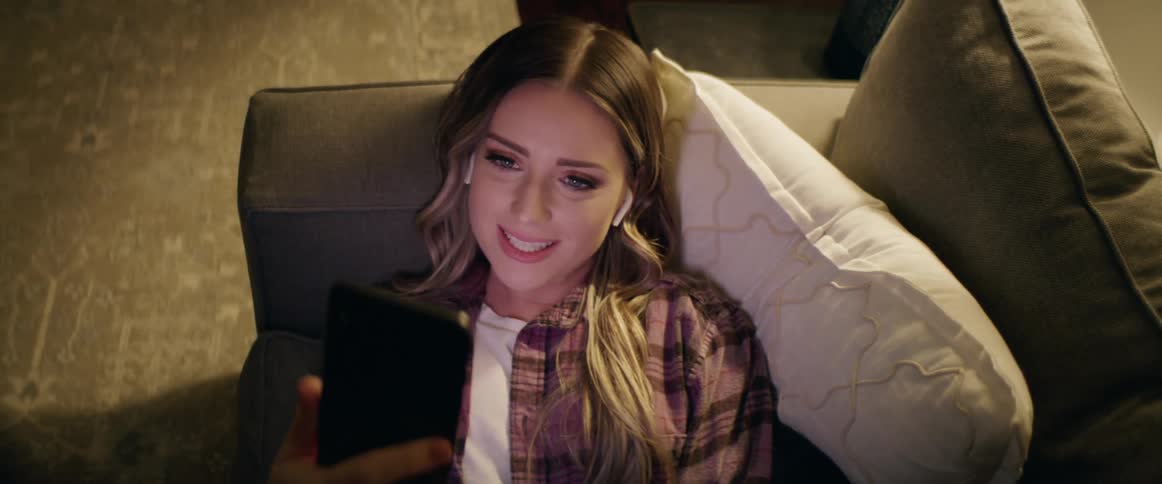 Female smiles and waves as she holds her phone out in front to video chat while laying on the sofa
