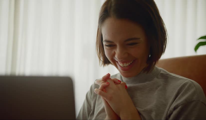 A woman claps her hands and laughs as she talks on a video call on her laptop at home