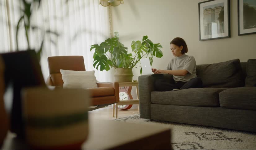 Female sitting on couch with her legs crossed underneath her types rapidly on a laptop that is resting on the arm of the couch