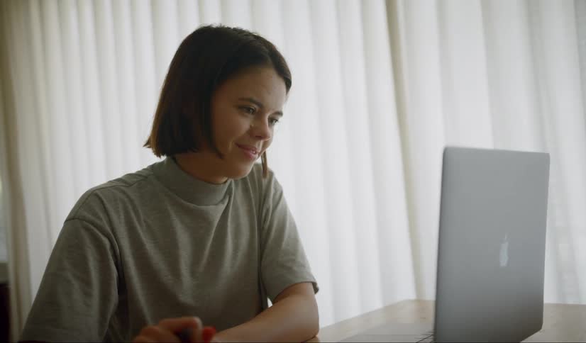 A teenager writes in a notebook and then smiles as she looks at her laptop