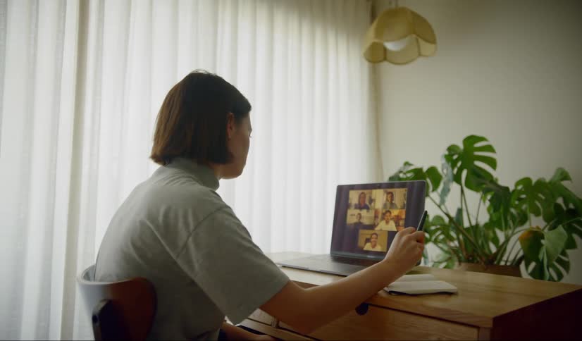A woman sits at a table and has a video meeting on a laptop