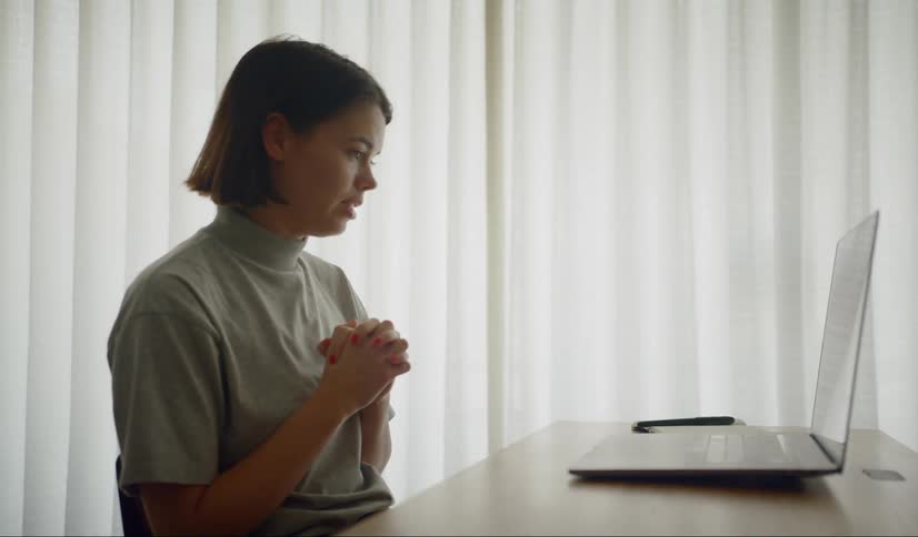 A woman sits at a table having a video meeting on a laptop