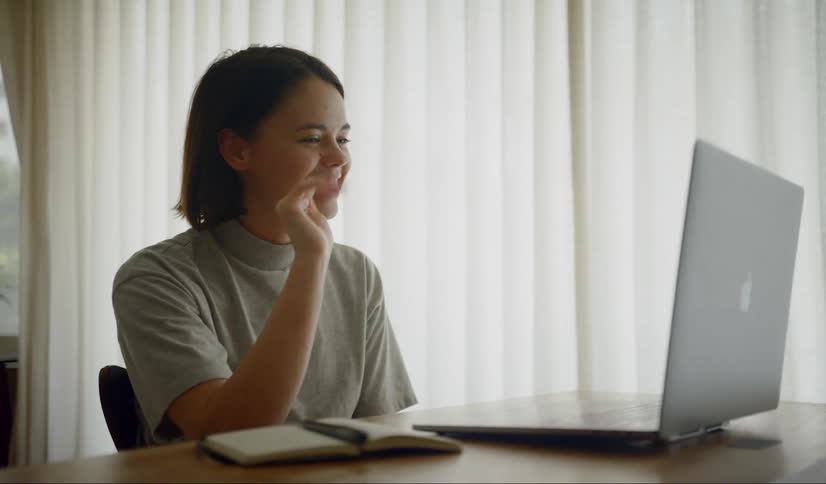 A woman sitting at a table video chatting and waving on a laptop