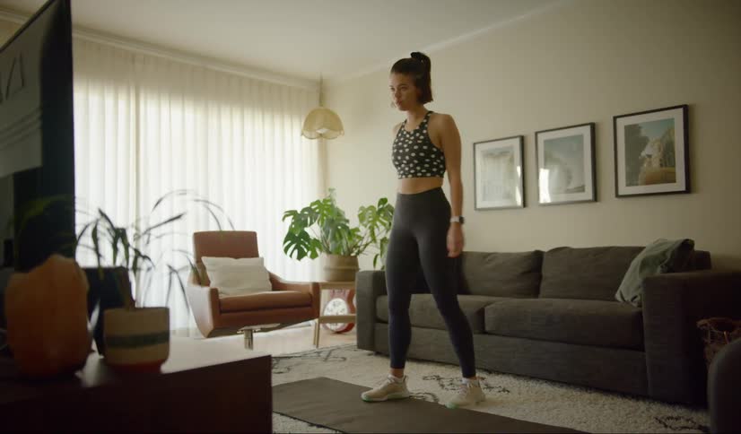 A woman stands on an exercise mat in her home facing her tv