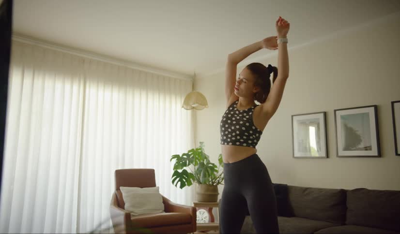 A woman in workout gear stretches with arms over her head standing in her living room
