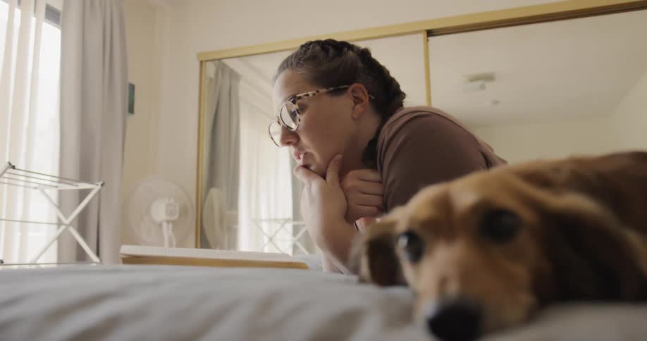 Female laying on her stomach on her bed with her chin propped up on her hand is reading a book while her dachshund lays next to her