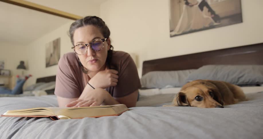 Woman reading book on bed while her dog is relaxing next to her