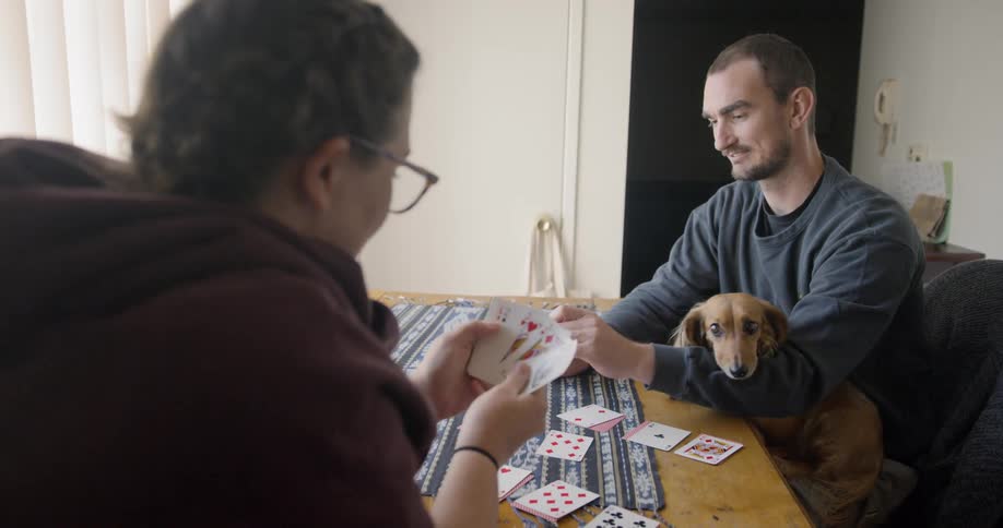 A man with a dog on his lap plays cards with a woman