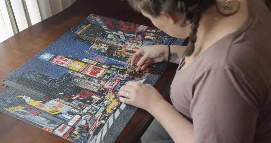 A woman sits at a table in front of a window finishing a jigsaw puzzle of times square