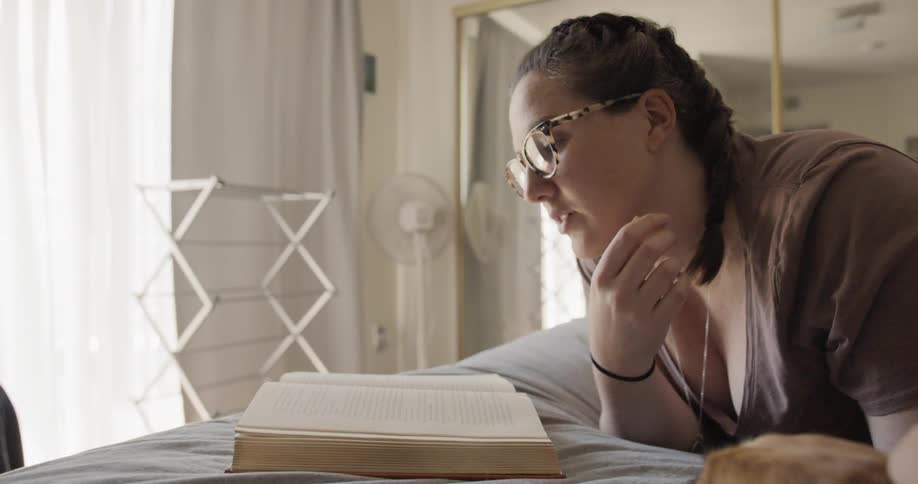 A woman lies on her bed reading a book tugging on her braid and petting her dog