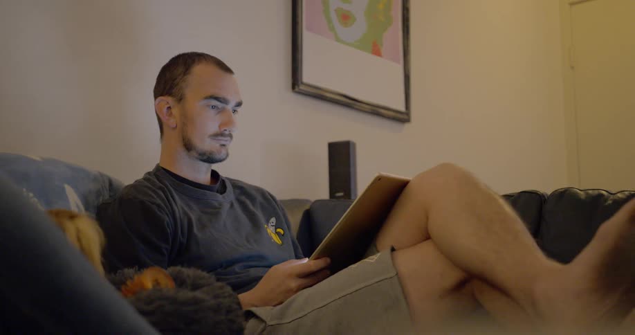 A man sits on his couch holding a tablet with his small dog in a dog bed beside him