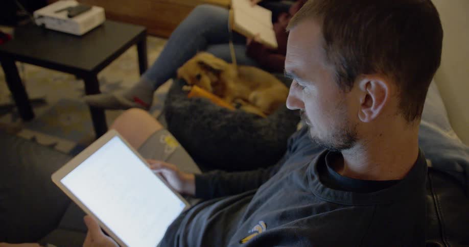A man sits on a couch holding a tablet while his wife reads a book and their dog plays with a toy
