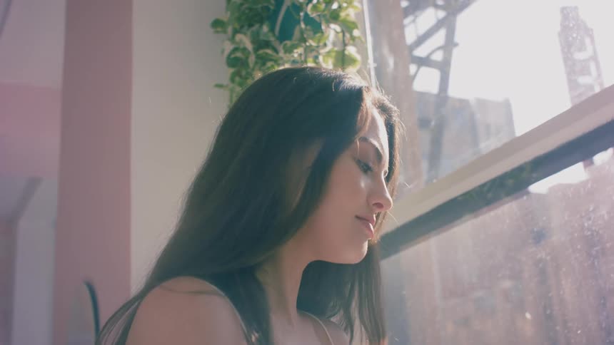 A woman flips her hair and looks out her sunny apartment window