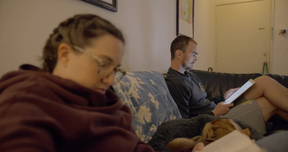 A man and woman sit on the couch reading with a dog in between them