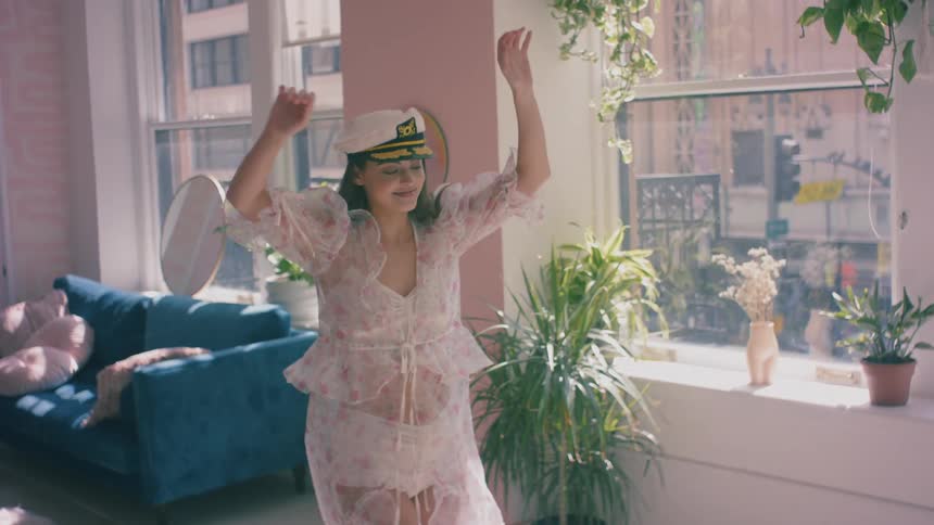A woman wearing lingerie and a sailing cap dances in her sunny city apartment