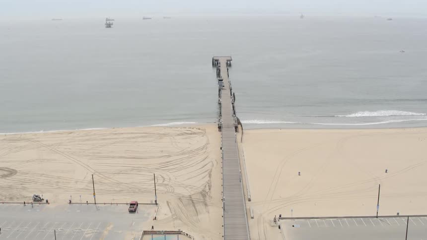 Aerial of a closed pier at the beach with a few people riding bikes in parking lot