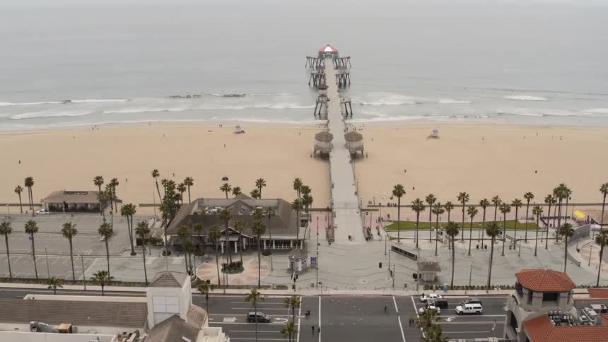 Cars cross intersection near buildings and pier leading across the beach into the ocean