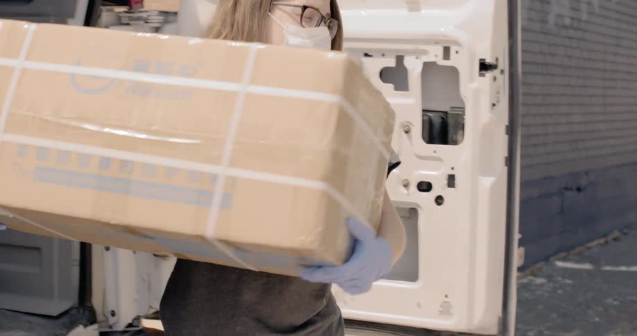 Woman wearing face mask unload cardboard boxes from van in alley at day