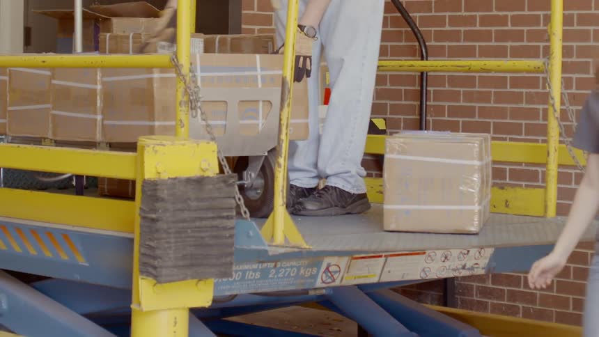 A woman unloads and places boxes on a lift where a man picks them up and stacks them on a dolly