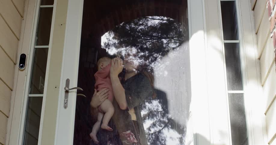 Woman holding her baby stands behind the glass door of the house