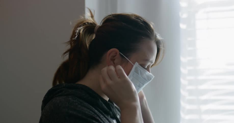 Woman puts on mask while standing near window