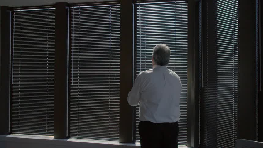 A man walks to an office window and opens the mini blinds letting light in
