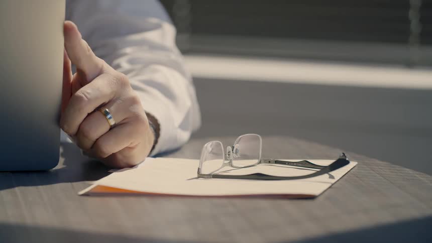A man in a white coat using a laptop takes his sets his glasses next to him on the desk