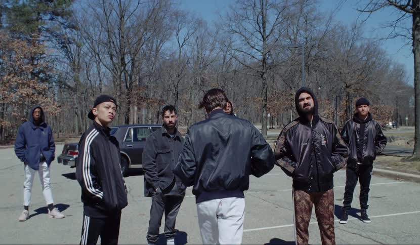 A group of male teens stand outside and look at something in the distance and slowly walk forwards