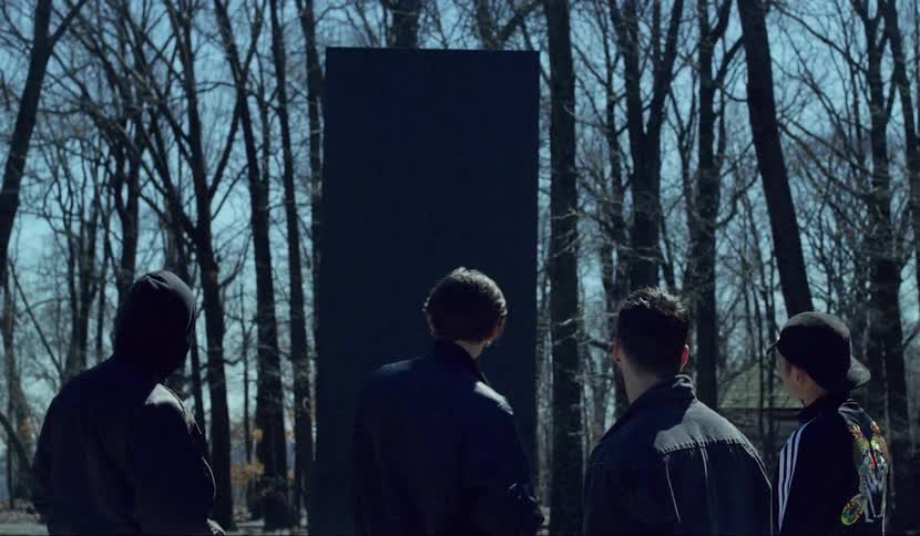 A young man coming to stand with other young men in front of a black monolith in a woodland