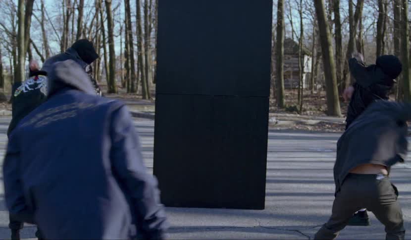 A group of men perform a choreographed dance in the parking of a public park near a forest