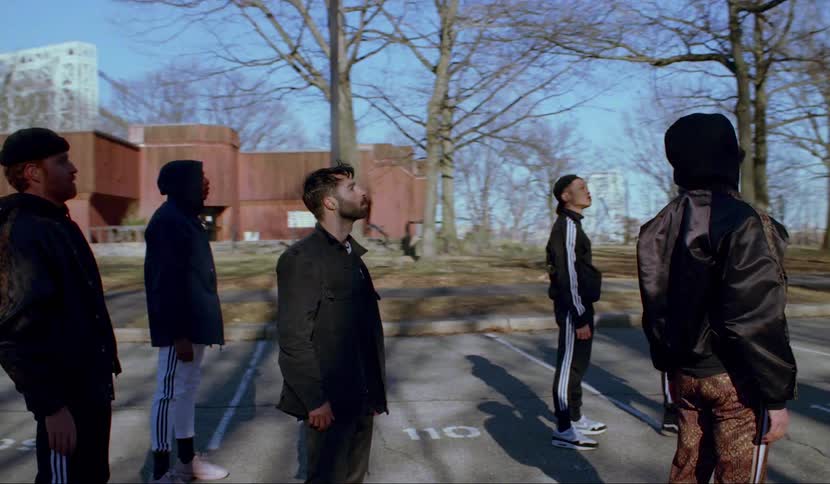A group of men stand in a parking lot on a sunny day and perform head tilts in a choreographed manner