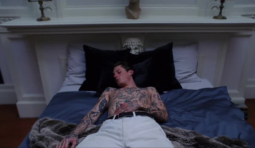 A shirtless man with tattoos covering his chest and arms is sleeping on top of blankets on a bed