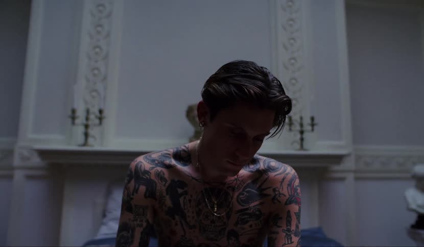 A shirtless and tattooed man stands in the front of a church with his head bowed