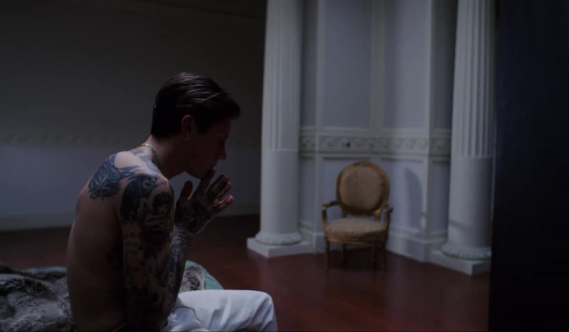 A shirtless man with tattoos sits on a bed nervously rubbing and clasping his hands while looking at a large black panel