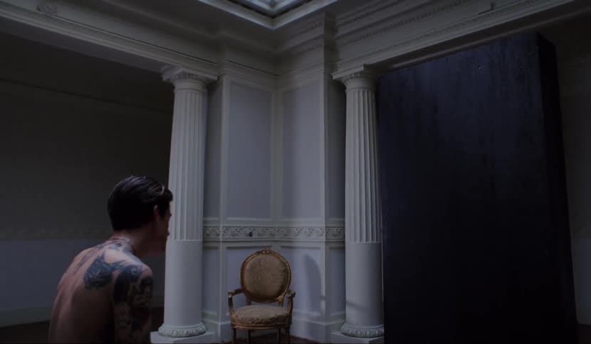A shirtless man with tattoos sits on the end of a bed looking at a large black panel