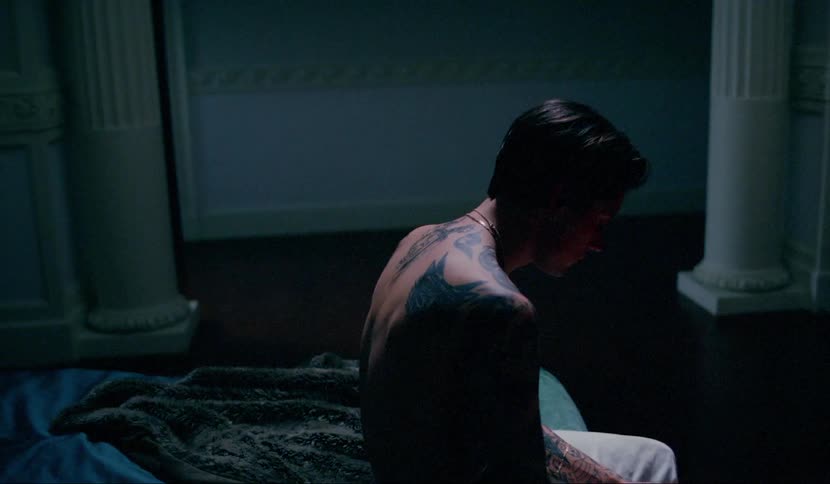 A tattooed shirtless man sits on the edge of a bed