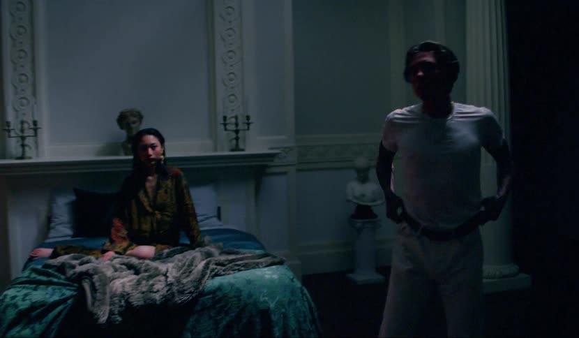 A woman sits on a bed and watches a man stagger to his feet from the floor then pace and talk