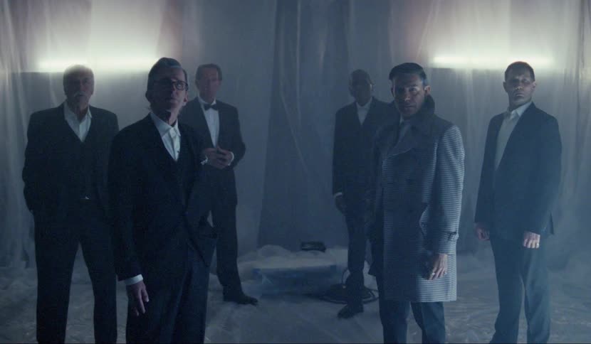 A group of older men wearing formal attire stand in a room covered in plastic tarps staring