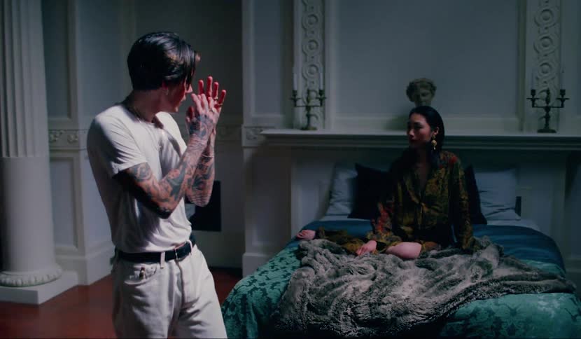A woman sits on a bed listening and watching as a man paces and gestures and talks in an anxious manic manner