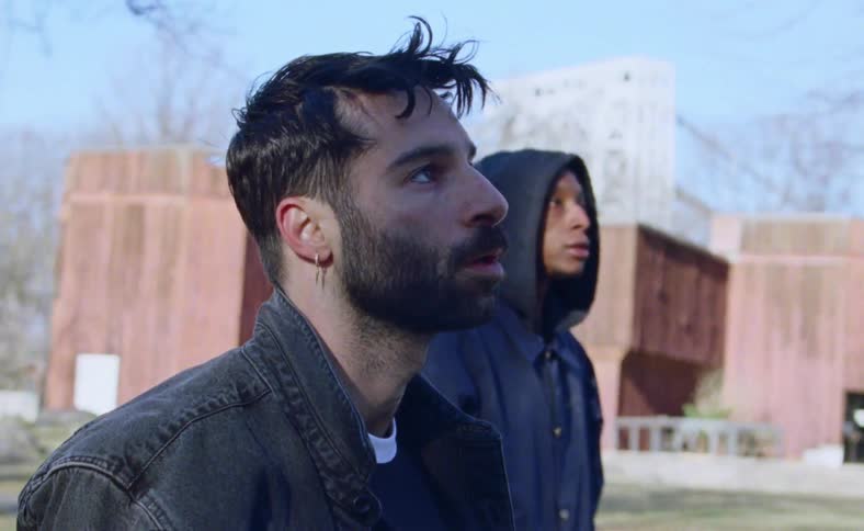 Men stand frozen staring nervously among leafless trees with a building in the background