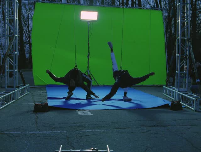 Two men hanging by wires in front of a green screen and a light