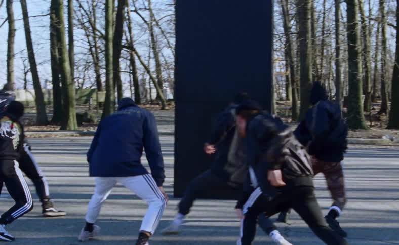 A group of men dance choreographed steps in front of a large black panel in a parking lot on a sunny day