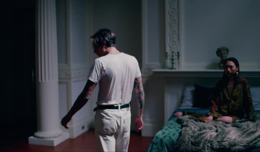 An asian woman sits on a bed as a young man walks around the room talking and then drops to the floor and puts his head in his hands