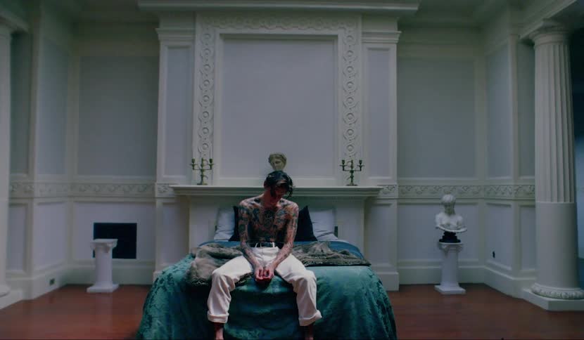 A young male covered in tattoos sits on a bed with his head hung down