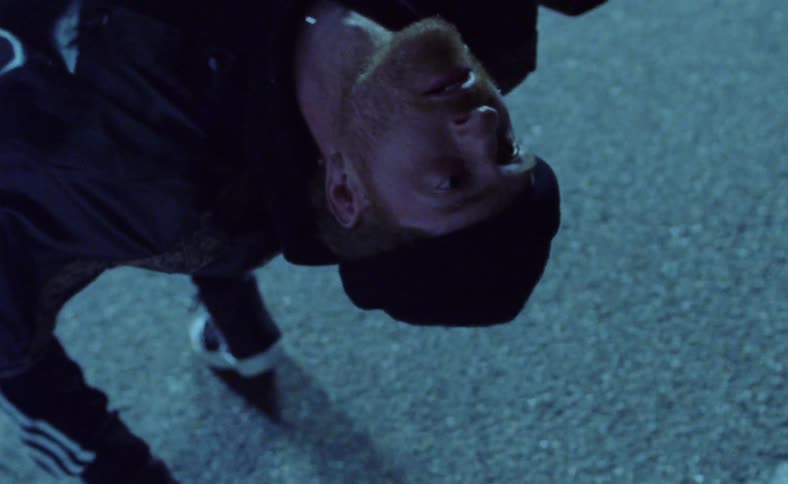 A man facing a large glowing screen in a parking lot falls backwards suspended by wire