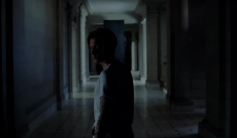 Man walks down a dark hallway and turns to look behind him then keeps walking