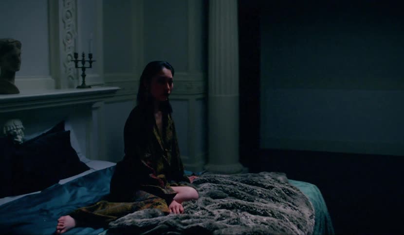 An asian woman sitting on a bed looking at the camera as it circles her