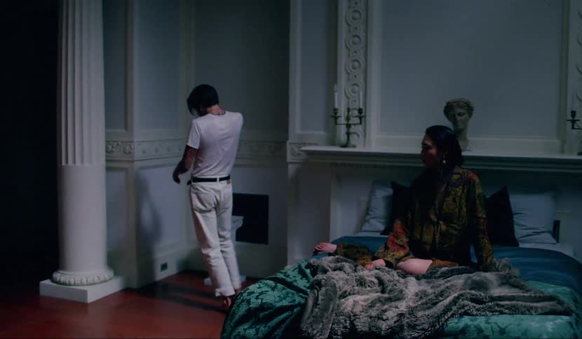 A man paces haphazardly around an ornate room as a woman sits atop a bed watching him