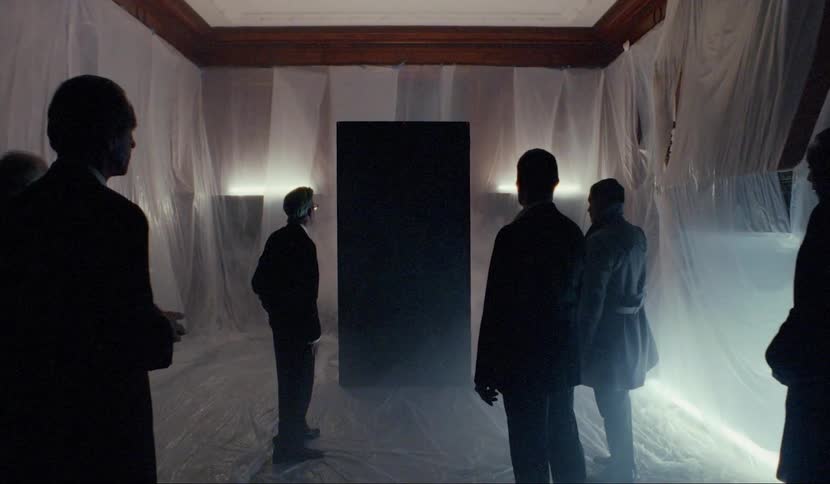 A group of men standing in front of a black monolith in a room covered in plastic